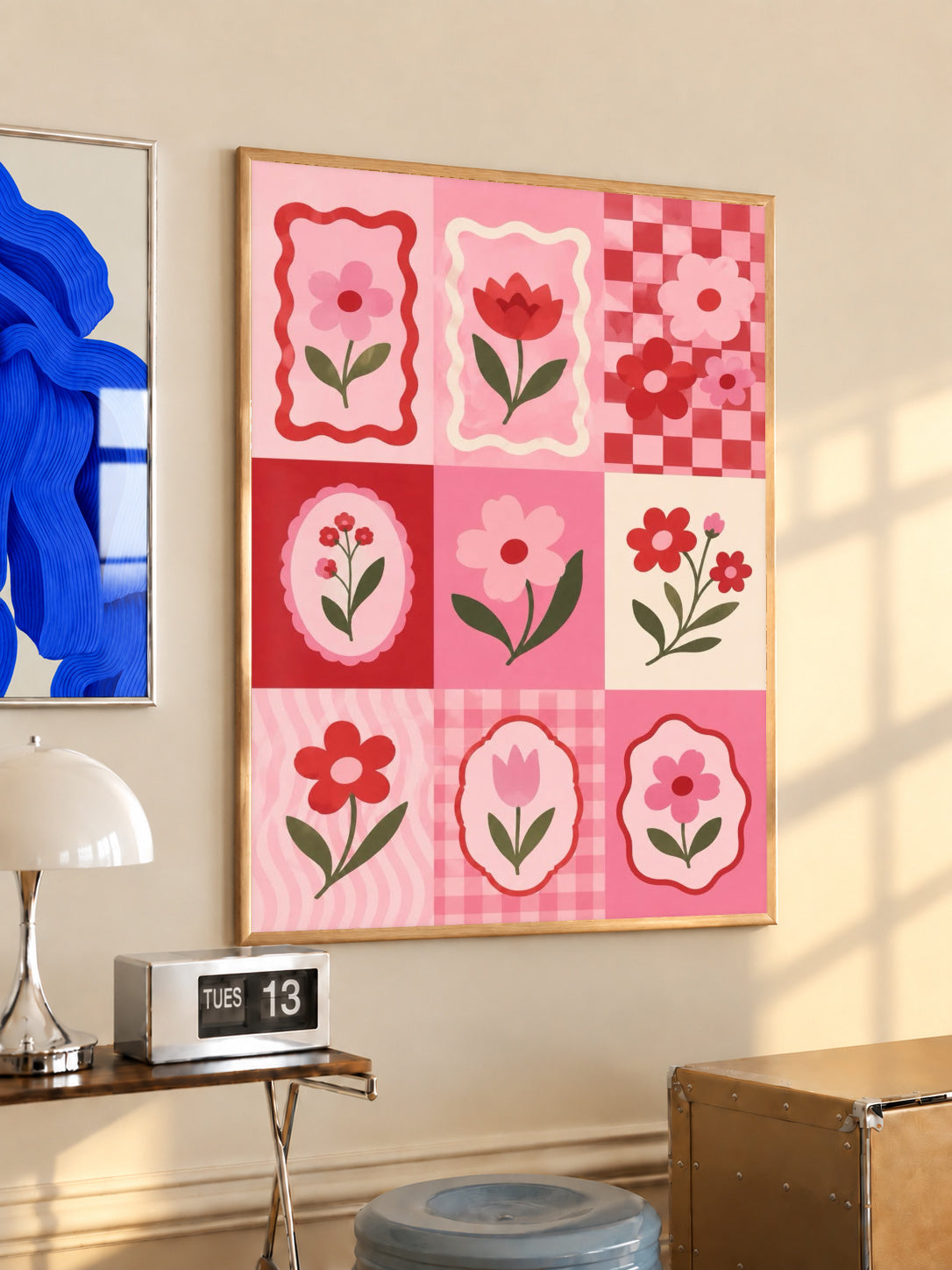 A pink and red patchwork grid of tulips, daisies, scalloped frames and checkerboard squares in a light wood frame on a sunlit office wall beside a desk