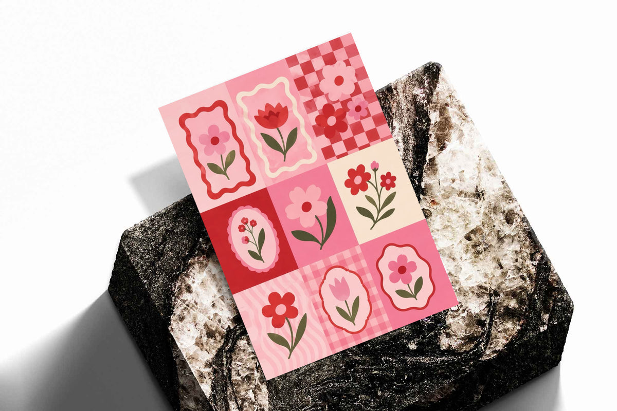 A pink and red patchwork grid of tulips, daisies, scalloped frames and checkerboard squares as an unframed print resting at an angle on a dark stone display