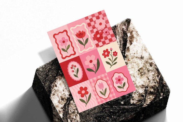 A pink and red patchwork grid of tulips, daisies, scalloped frames and checkerboard squares as an unframed print resting at an angle on a dark stone display