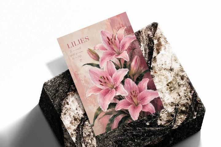 Pink lilies and buds with dark green leaves beneath a LILIES heading on a soft blush ground as an unframed print resting at an angle on a dark stone display