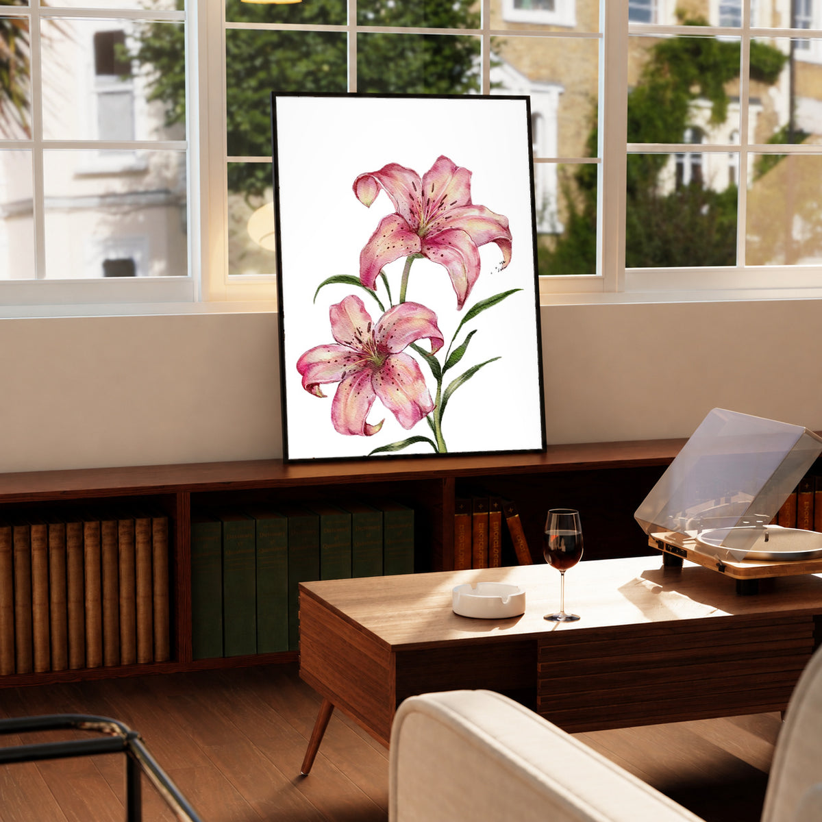 Two pink lilies with speckled petals and green leaves against white in a black frame on a wooden cabinet beside a window and record player