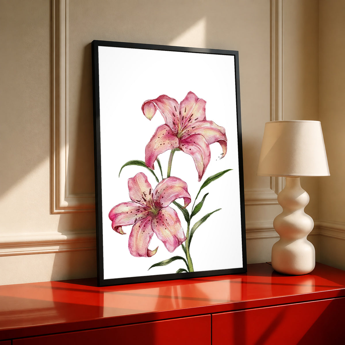 Two pink lilies with speckled petals and green leaves against white in a black frame on a red cabinet beside a cream lamp