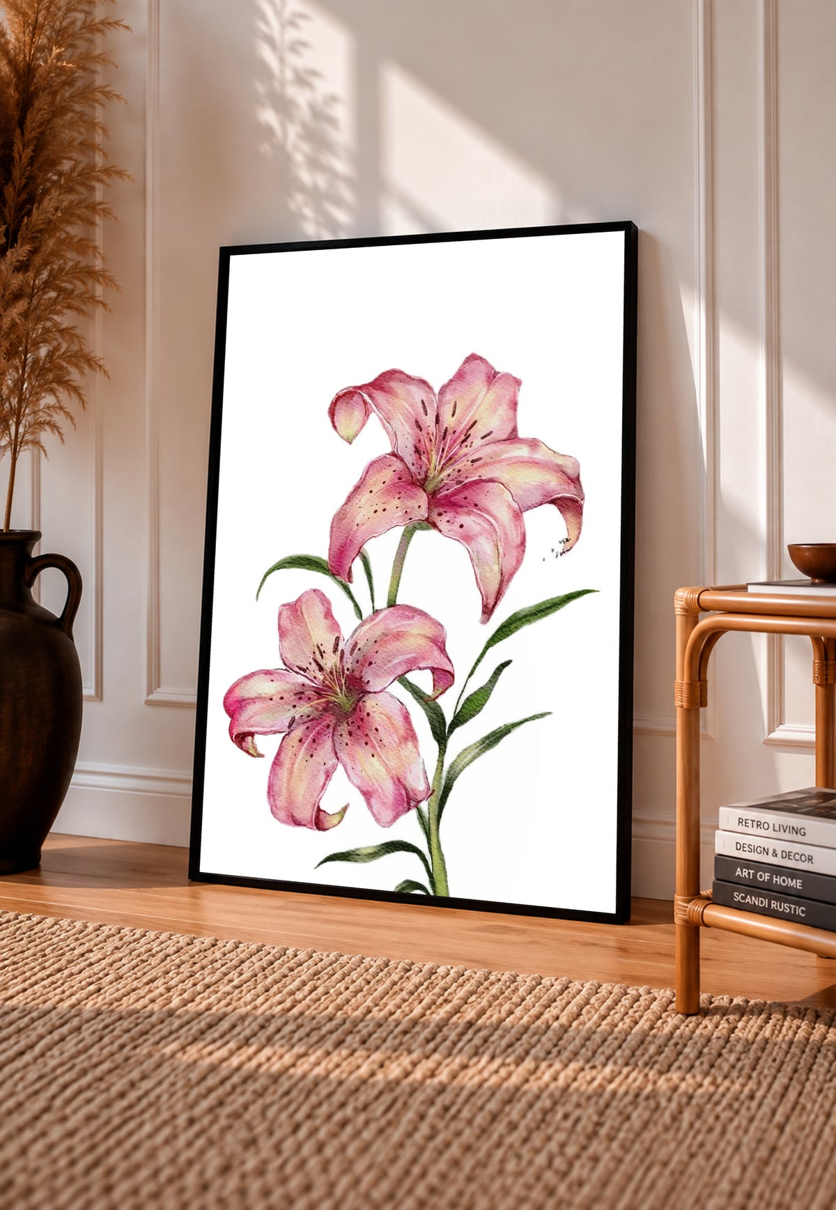 Two pink lilies with speckled petals and green leaves against white in a black frame leaning on a sunlit wooden floor beside books and a rattan seat