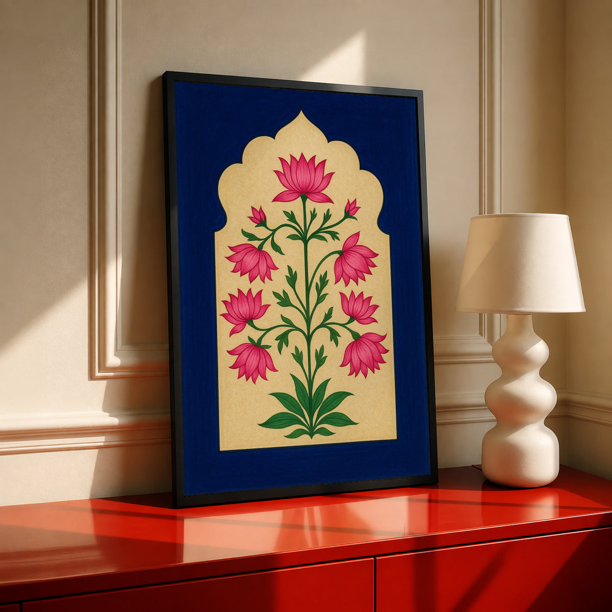 Pink lotus-like flowers and green stems within a scalloped gold arch on navy blue in a black frame on a red console beside a sculptural cream lamp