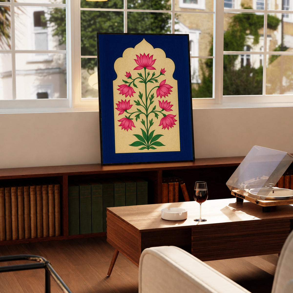 Pink lotus-like flowers and green stems within a scalloped gold arch on navy blue in a black display frame on a wooden console beside a sunlit window