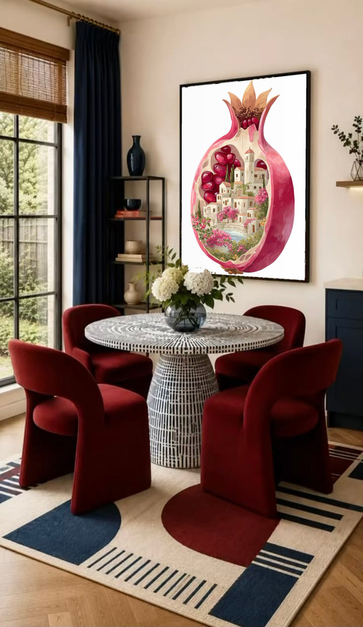 A cutaway pink pomegranate containing a tiny white village, ruby seeds and flowers in a black frame on the wall of a dining nook with red chairs