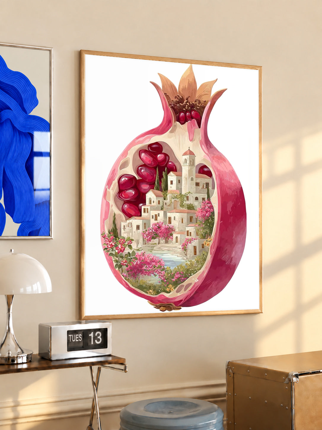 A cutaway pink pomegranate containing a tiny white village, ruby seeds and flowers in a light wood frame on a sunlit office wall beside a desk