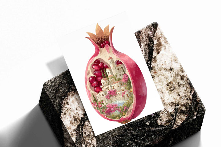 A cutaway pink pomegranate containing a tiny white village, ruby seeds and flowers as an unframed print resting at an angle on a dark stone display