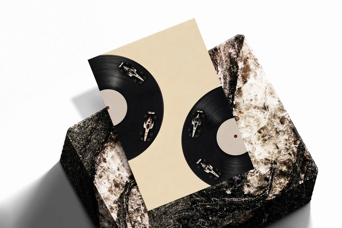 Four miniature race cars on the grooves of two black vinyl records against a cream background, shown as an unframed print tilted on a dark stone display against a light background