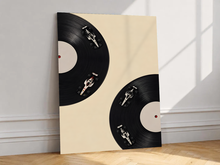 Four miniature race cars on the grooves of two black vinyl records against a cream background, shown unframed on a white wall above parquet flooring with a diagonal sun shadow