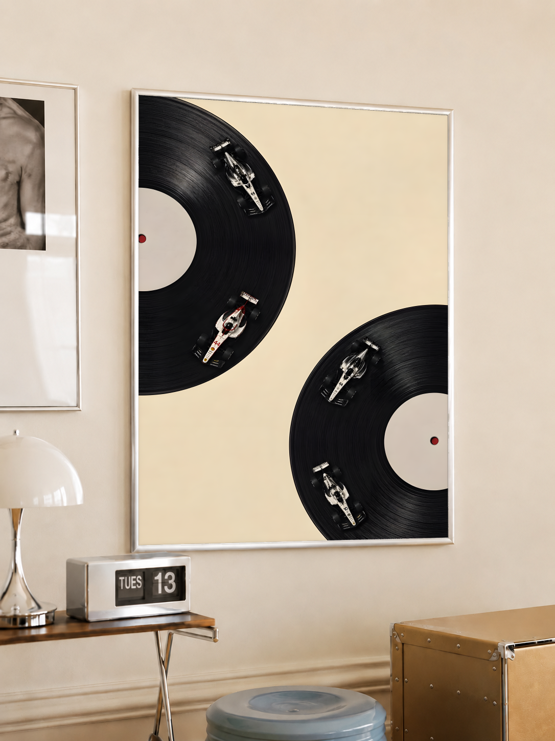 Four miniature race cars on the grooves of two black vinyl records against a cream background, styled in a light frame on a cream wall beside a white dome lamp and glass side table