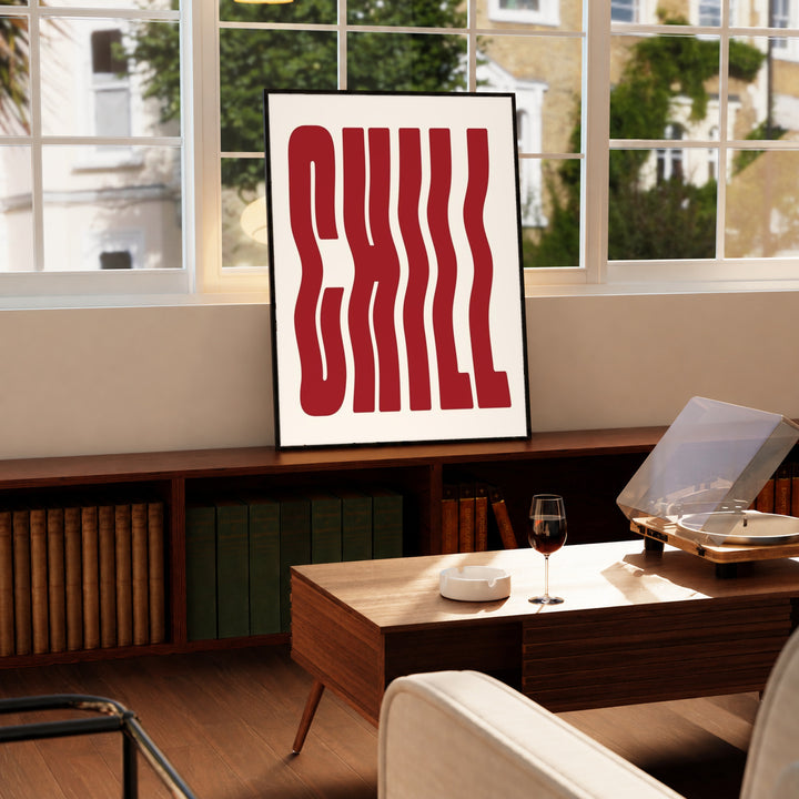 The word CHILL in tall wavy red lettering on a cream background in a black display frame on a wooden console beside a sunlit window