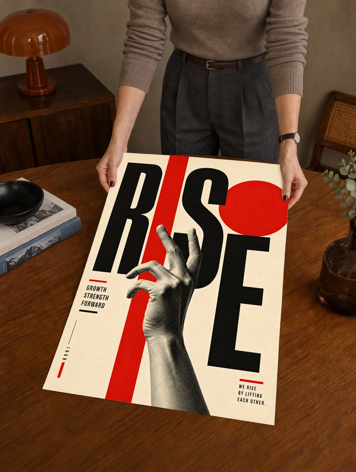 A reaching monochrome hand across oversized Rise lettering and red geometric shapes as a large unframed poster held above a wooden desk