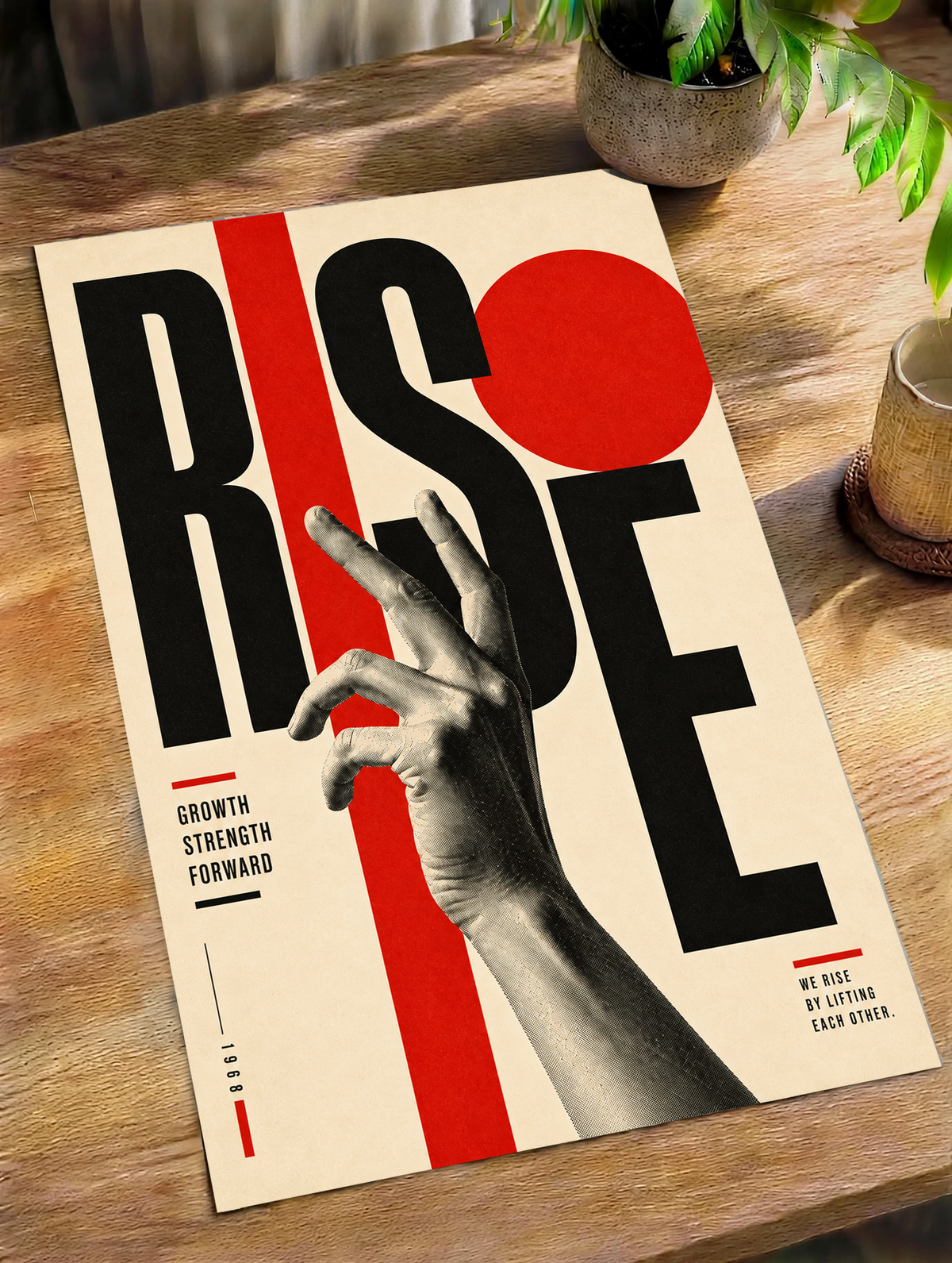 A reaching monochrome hand across oversized Rise lettering and red geometric shapes as an unframed poster lying on a wooden table beside a plant