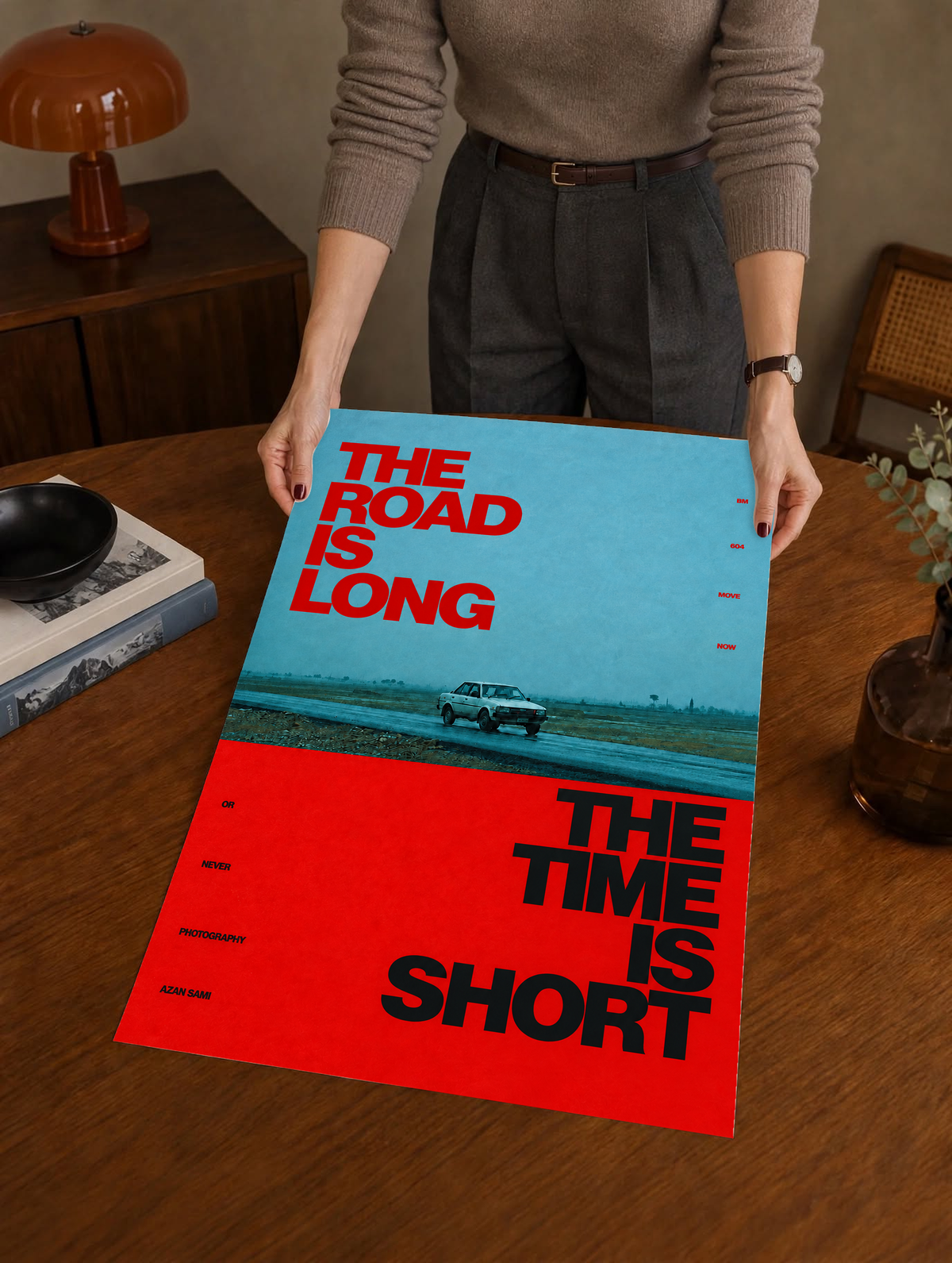 A small vintage car between blue and red blocks with bold road and time lettering as a large unframed poster held above a wooden desk