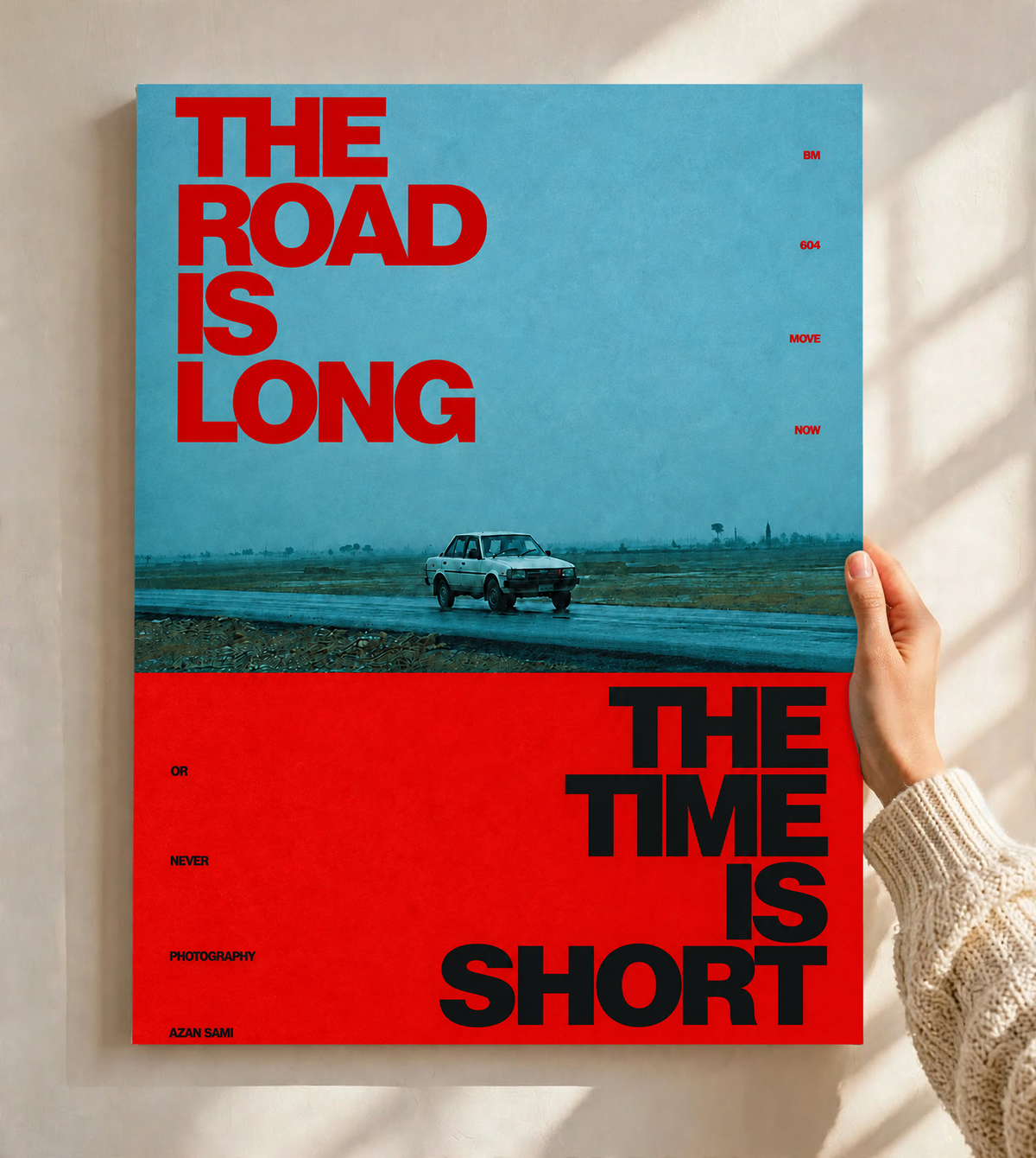 A small vintage car between blue and red blocks with bold road and time lettering as an unframed poster held against a pale sunlit wall