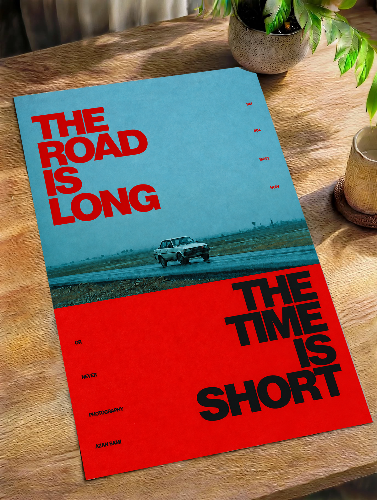 A small vintage car between blue and red blocks with bold road and time lettering as an unframed poster lying on a wooden table beside a plant