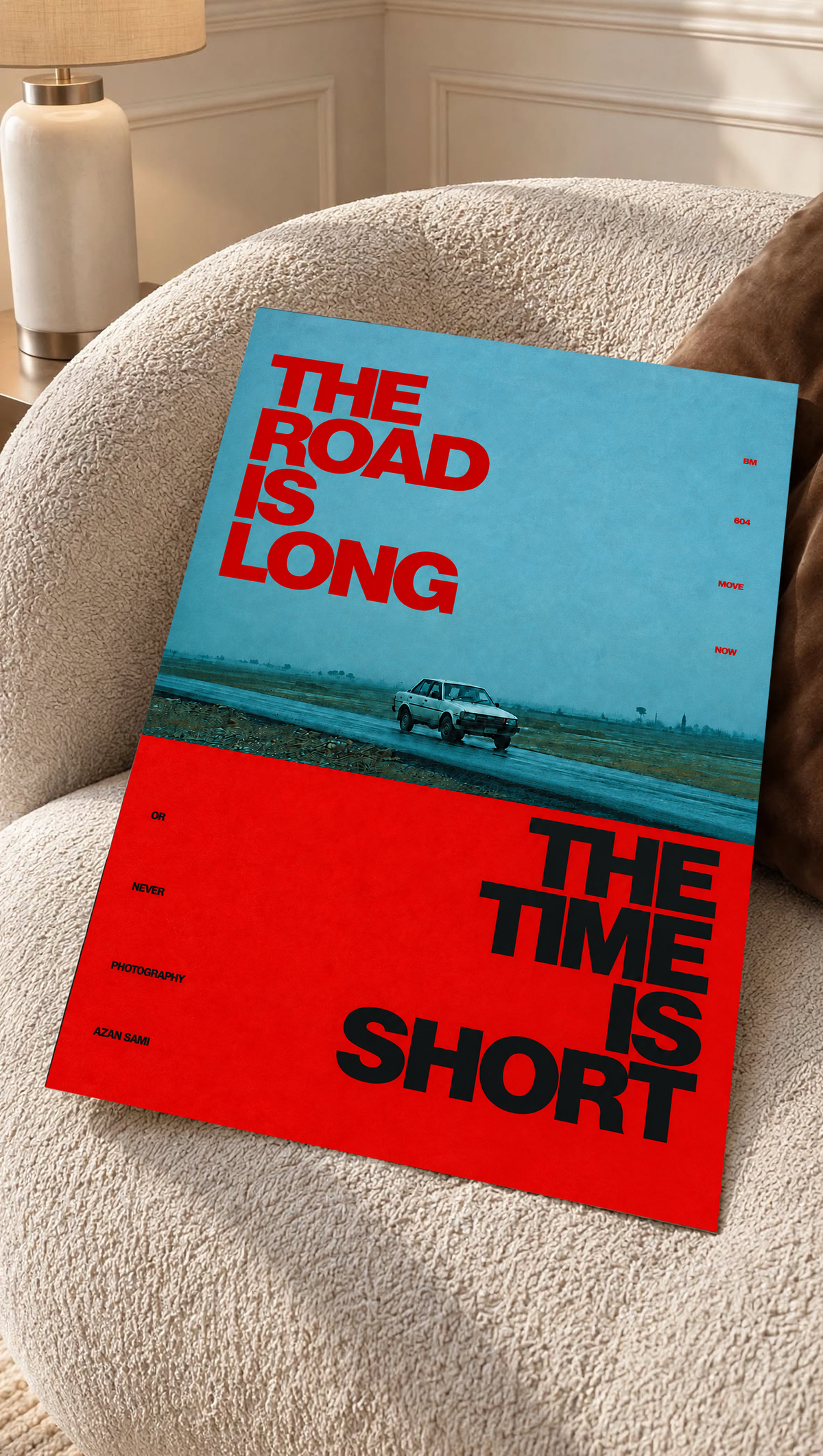 A small vintage car between blue and red blocks with bold road and time lettering as an unframed poster resting on a cream woven chair
