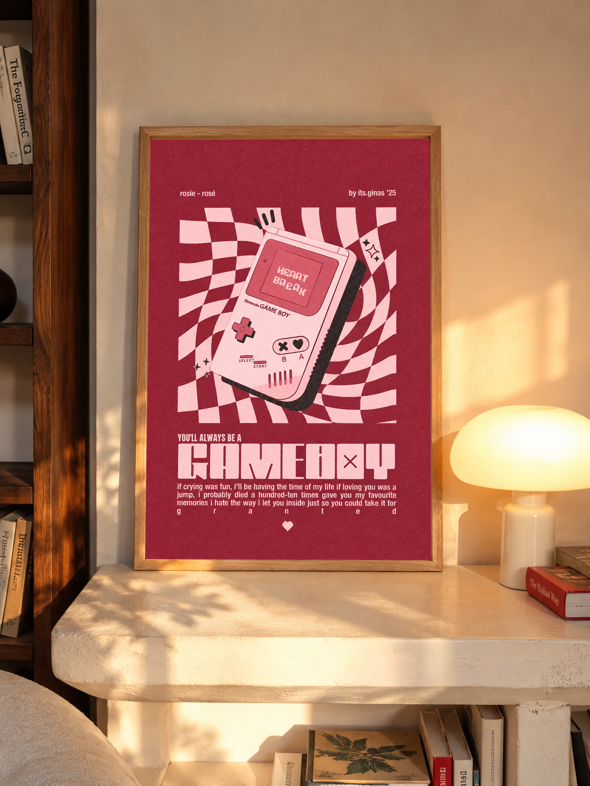 A pink Game Boy-style console with Heart Break screen on a wavy pink checkerboard in an oak frame above a pale wooden sideboard beside a glowing lamp