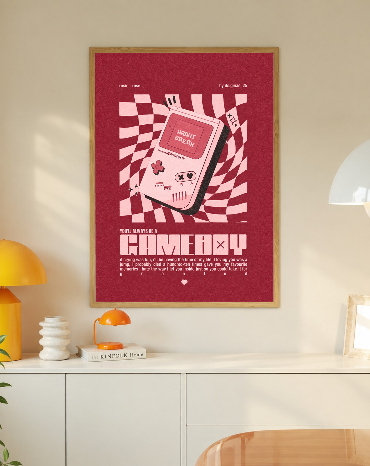 A pink Game Boy-style console with Heart Break screen on a wavy pink checkerboard in a slim oak frame on a cream wall above a white cabinet