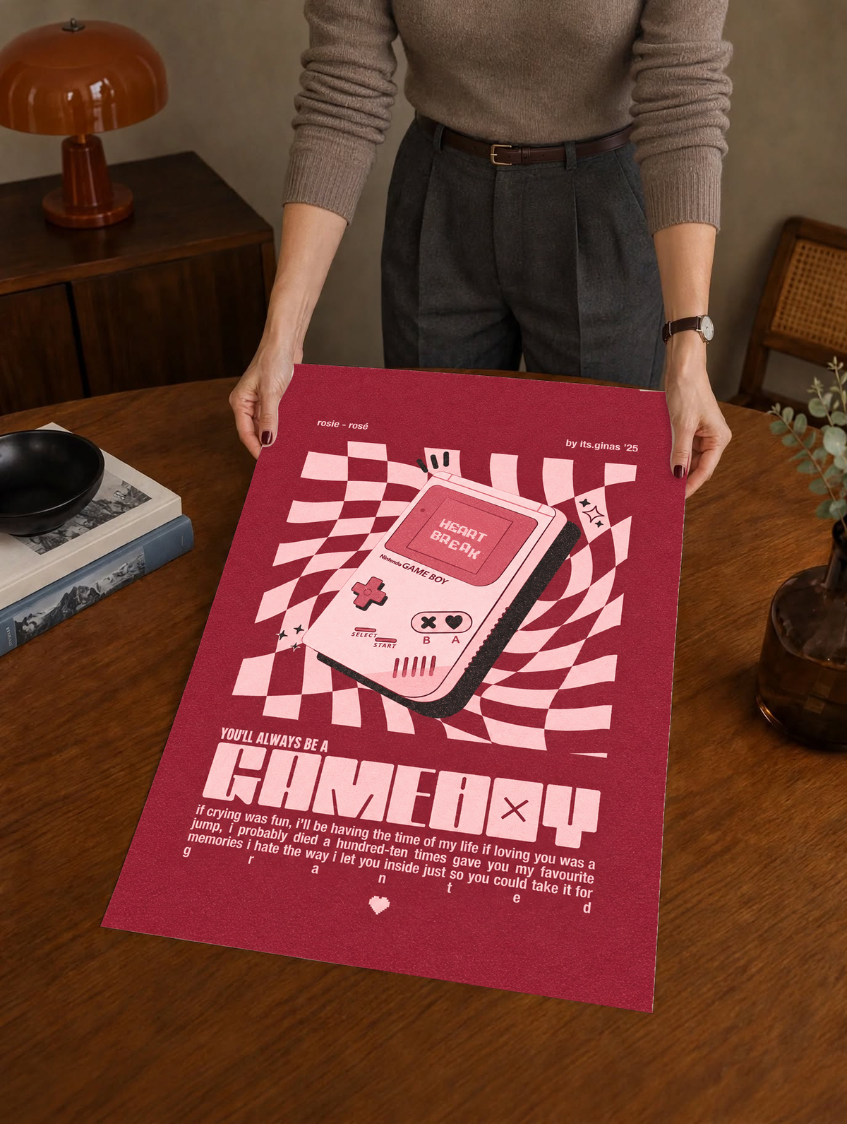 A pink Game Boy-style console with Heart Break screen on a wavy pink checkerboard as a large unframed poster held above a wooden desk