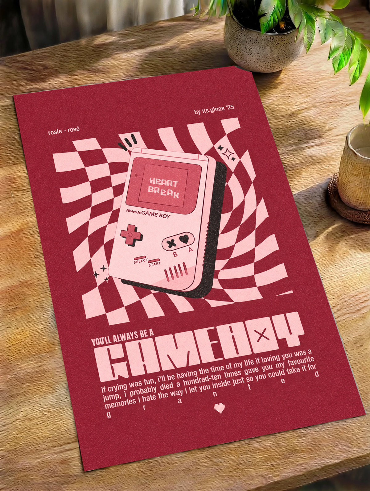 A pink Game Boy-style console with Heart Break screen on a wavy pink checkerboard as an unframed poster lying on a wooden table beside a plant