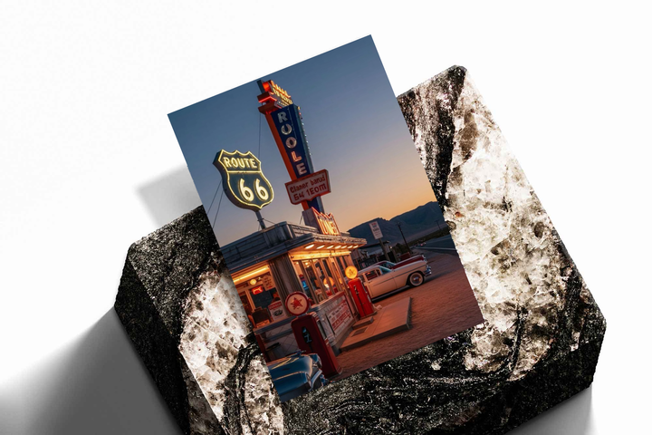 Unframed artwork depicting neon diner sign with cars and roadside pumps at dusk, presented diagonally on rough stone.