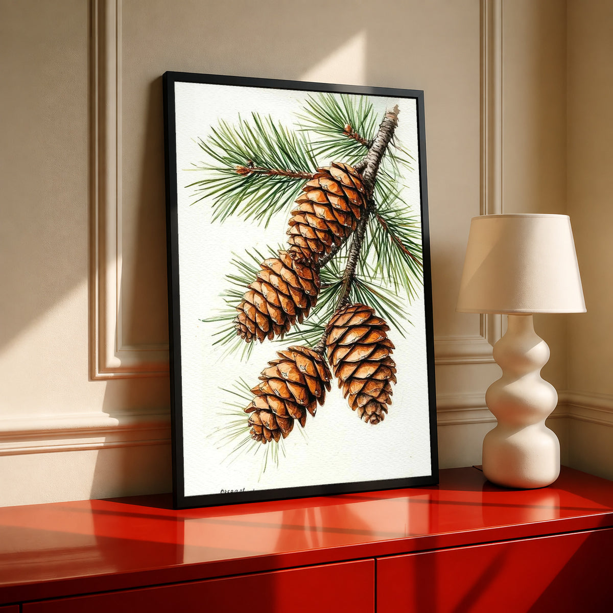 Three brown pine cones on a branch with long green needles against white in a black frame on a red cabinet beside a cream lamp