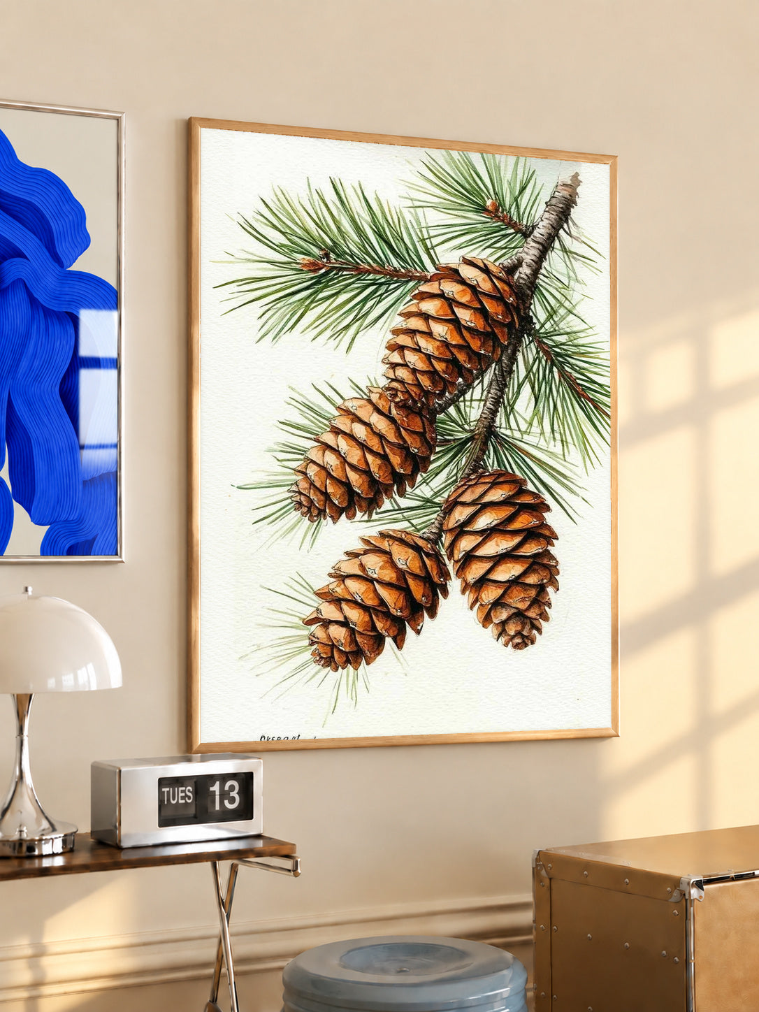 Three brown pine cones on a branch with long green needles against white in an oak frame above a console beside a blue print and calendar