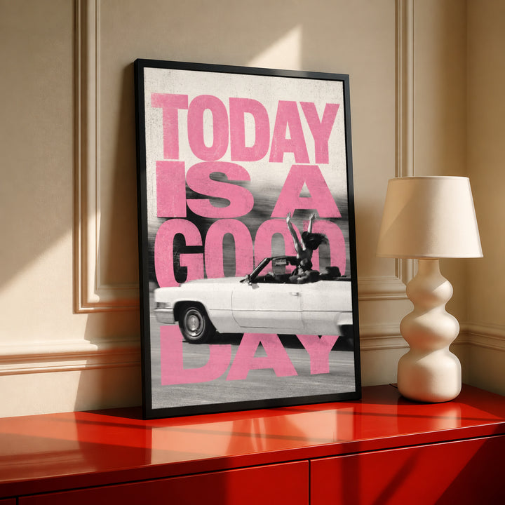 Bold pink TODAY IS A GOOD DAY lettering behind a black-and-white convertible car collage in a black frame on a red console beside a sculptural cream lamp