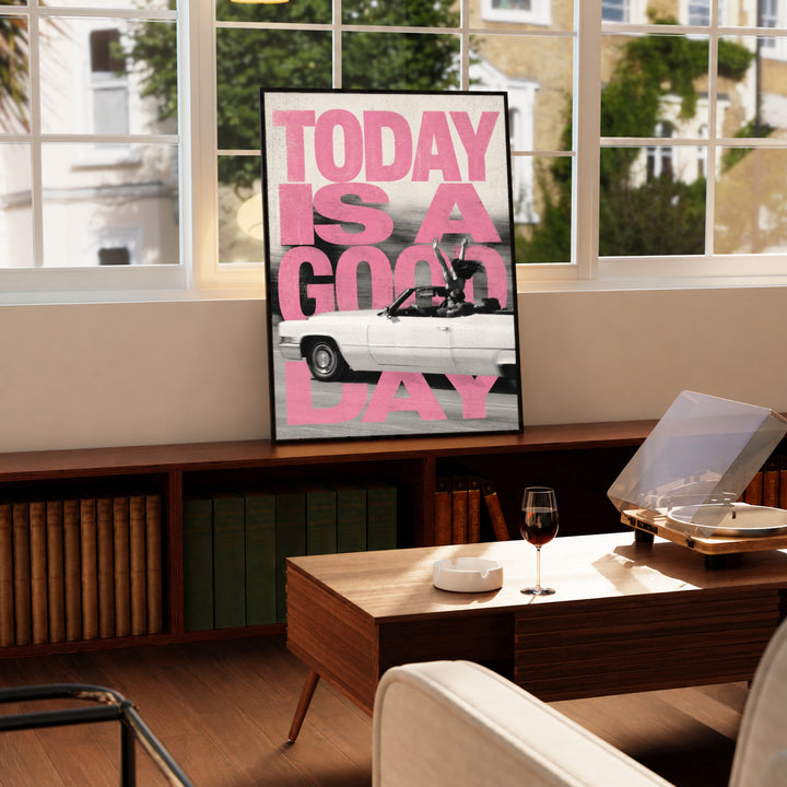 Bold pink TODAY IS A GOOD DAY lettering behind a black-and-white convertible car collage in a black display frame on a wooden console beside a sunlit window