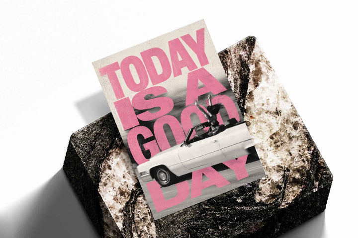 Bold pink TODAY IS A GOOD DAY lettering behind a black-and-white convertible car collage as an unframed print resting at an angle on a dark stone display