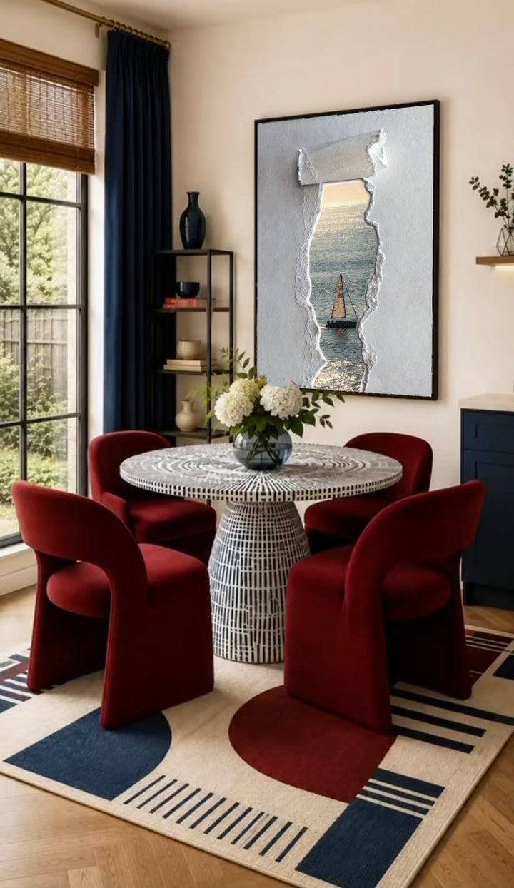 A sailboat at sea seen through a printed torn-paper opening on a grey ground in a black frame on the wall of a dining nook with red chairs