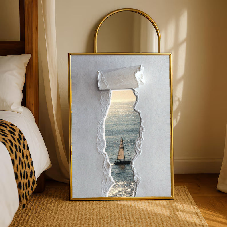 A sailboat at sea seen through a printed torn-paper opening on a grey ground in a gold-tone styling frame leaning beside a bedroom mirror