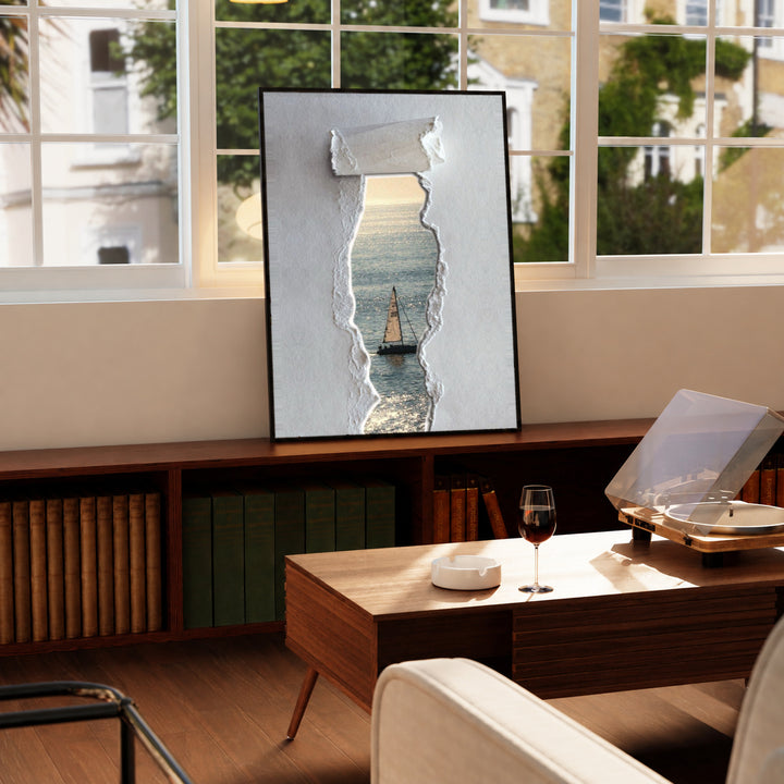 A sailboat at sea seen through a printed torn-paper opening on a grey ground in a black display frame on a wooden console beside a sunlit window