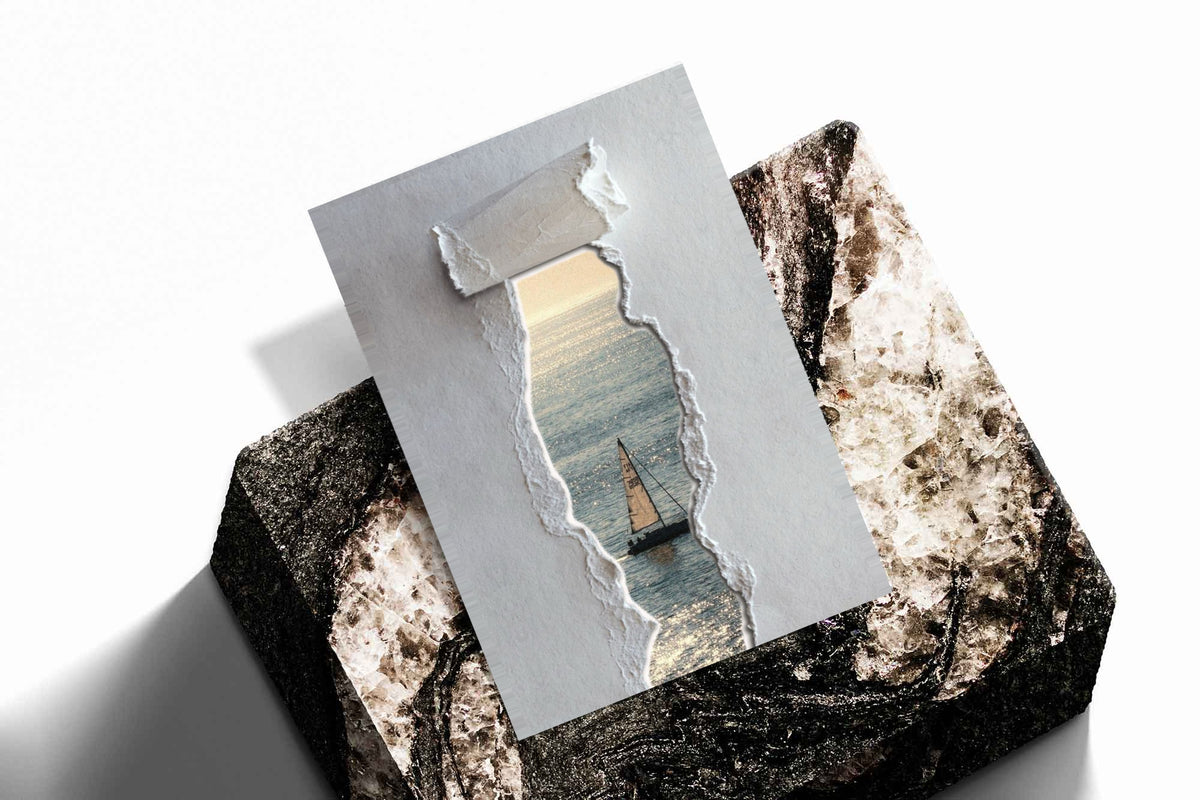 A sailboat at sea seen through a printed torn-paper opening on a grey ground as an unframed print resting at an angle on a dark stone display