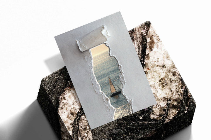 A sailboat at sea seen through a printed torn-paper opening on a grey ground as an unframed print resting at an angle on a dark stone display