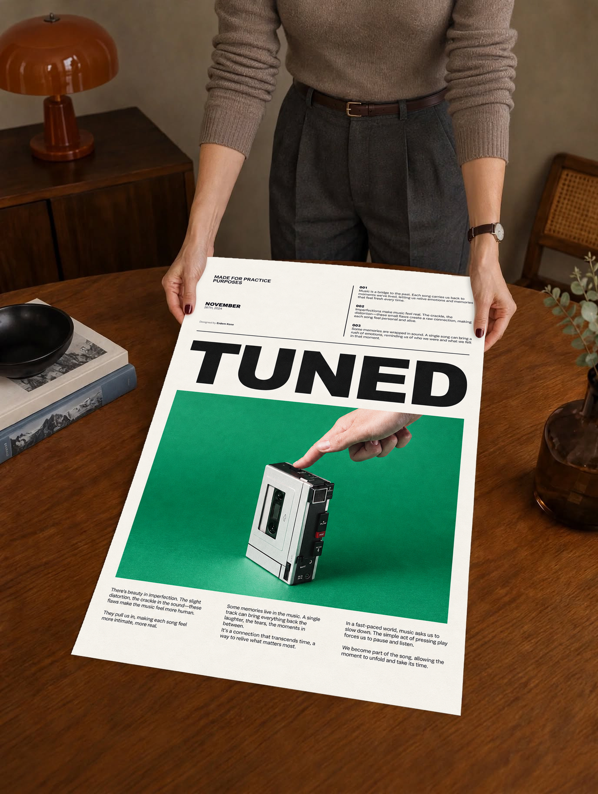 A hand pressing a portable cassette player against green beneath Tuned typography as a large unframed poster held above a wooden desk