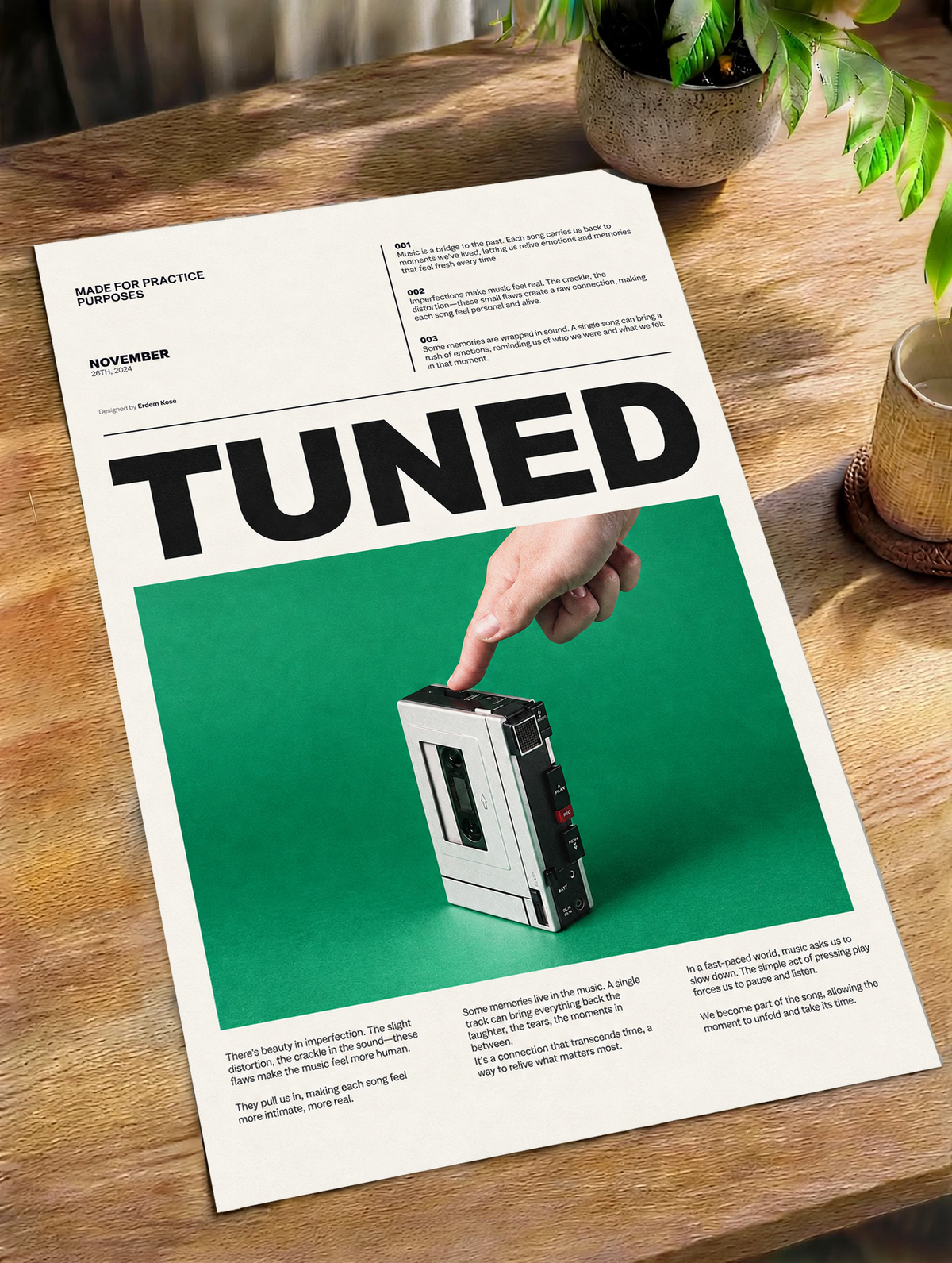 A hand pressing a portable cassette player against green beneath Tuned typography as an unframed poster lying on a wooden table beside a plant