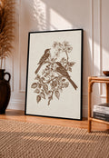 Two birds perched on a flowering branch with leaves and blossoms in sepia in a black frame leaning on a sunlit wooden floor beside books and a rattan seat