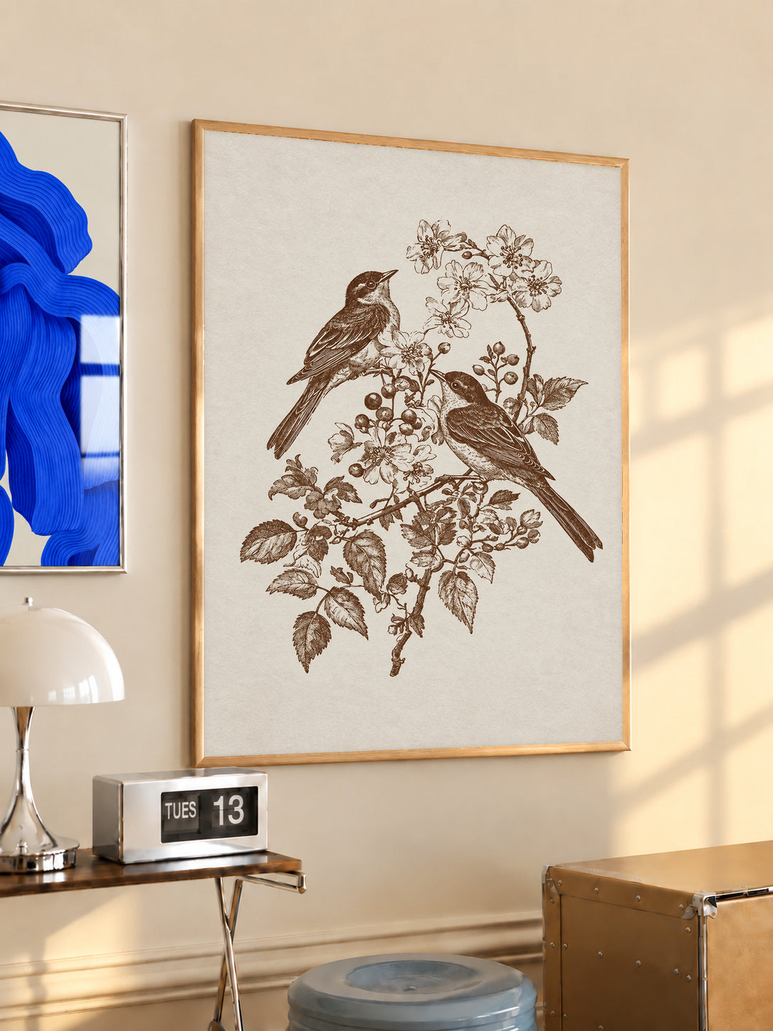 Two birds perched on a flowering branch with leaves and blossoms in sepia in an oak frame above a console beside a blue print and calendar