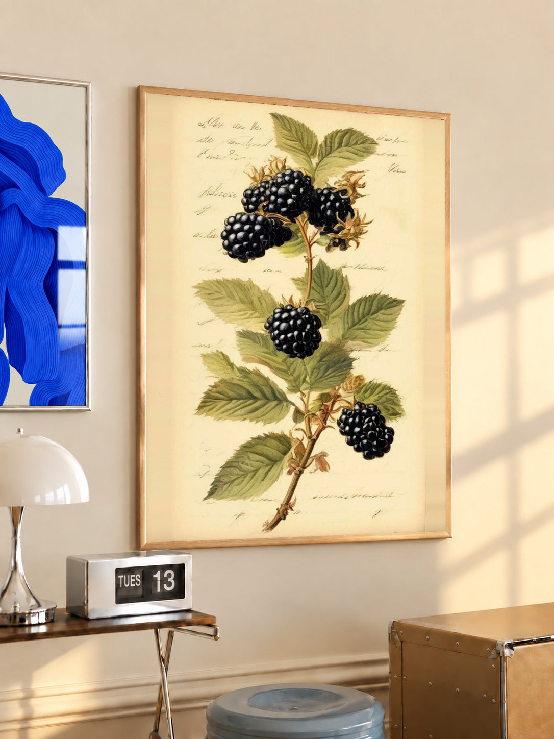 Dark blackberry clusters and serrated green leaves on a branch with notes on aged cream in an oak frame above a console beside a blue print and calendar