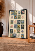 Grid of twenty four illustrated floral postage stamps in muted garden colours in a black frame leaning on a sunlit wooden floor beside books and a rattan seat