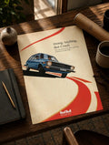 Lámina sobre una mesa de madera: coche retro azul sobre curvas rojas en fondo crema, con eslogan y texto Red Bull.