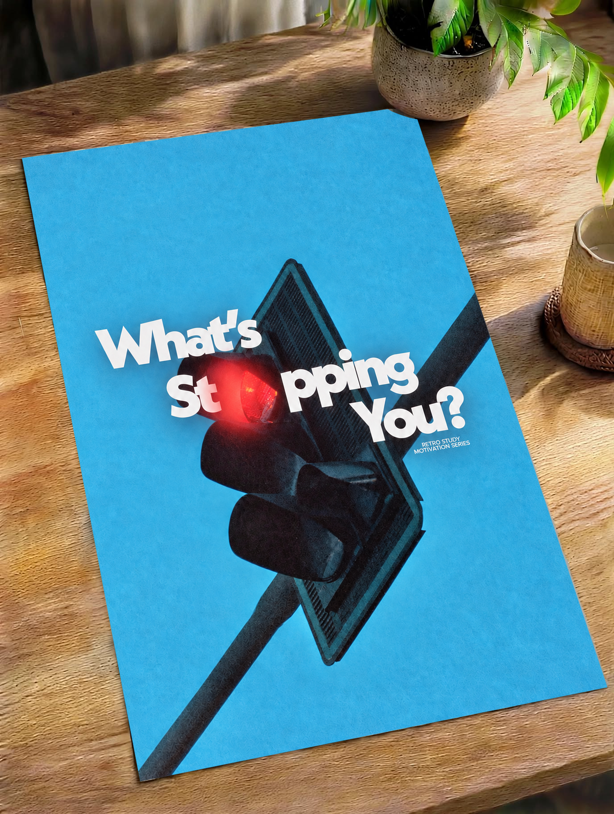White What's Stopping You lettering over a red traffic light against blue sky as an unframed poster lying on a wooden table beside a plant