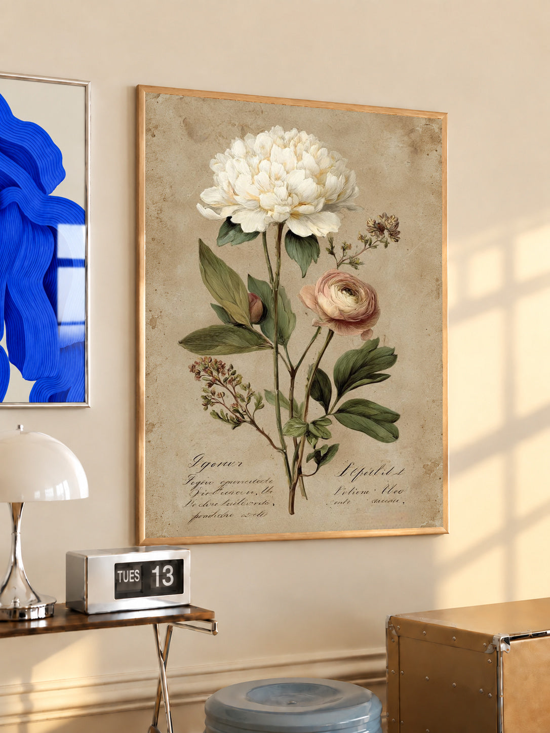 Large white peony and smaller pink layered flower with green leaves on aged cream in an oak frame above a console beside a blue print and calendar
