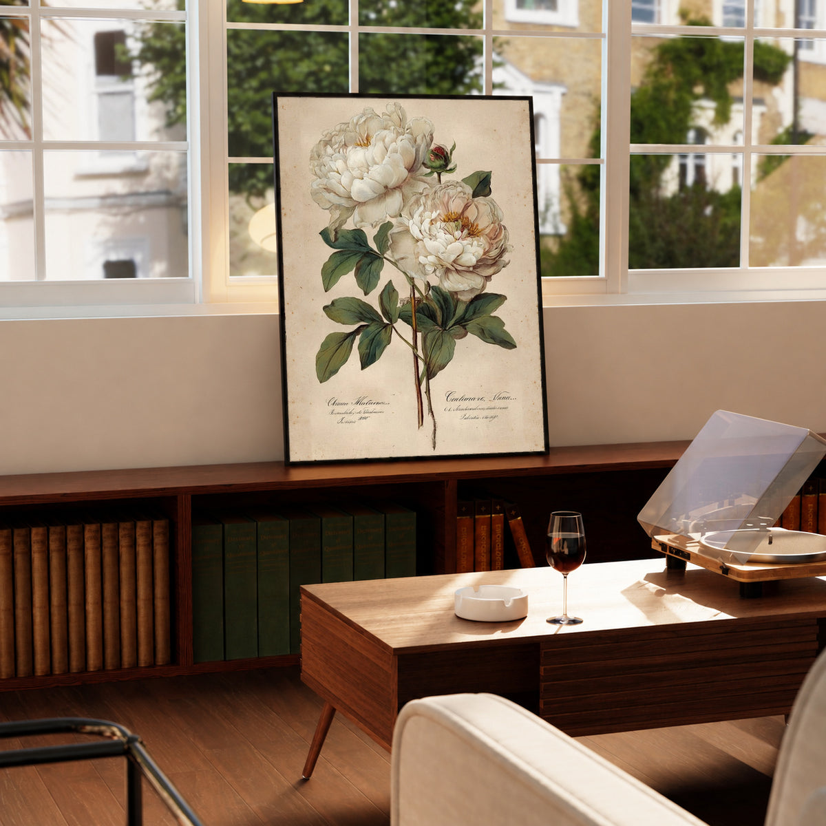 Two white peony blooms and a small bud with green leaves on aged cream in a black frame on a wooden cabinet beside a window and record player