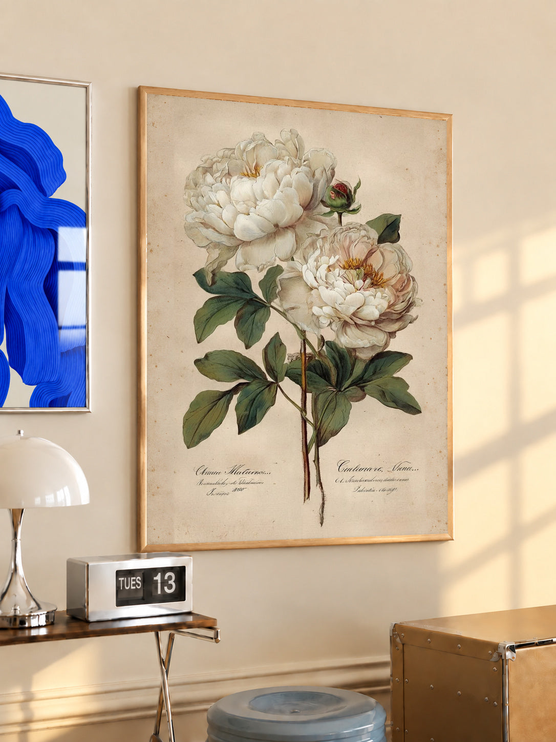 Two white peony blooms and a small bud with green leaves on aged cream in an oak frame above a console beside a blue print and calendar