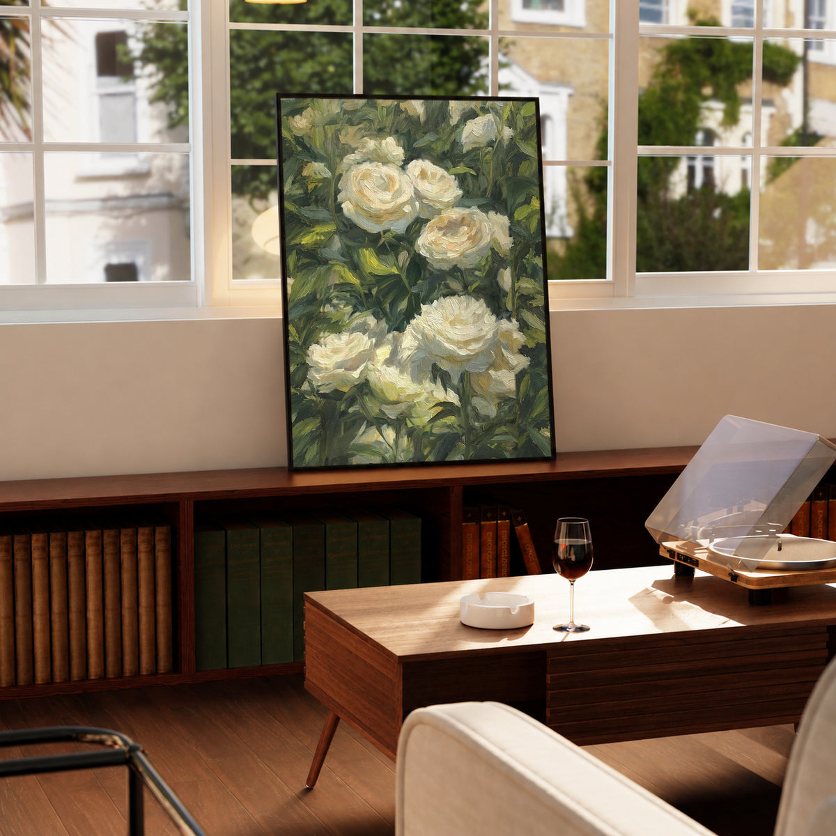 Cream white roses among dense green garden foliage in a painterly composition in a black frame on a wooden cabinet beside a window and record player