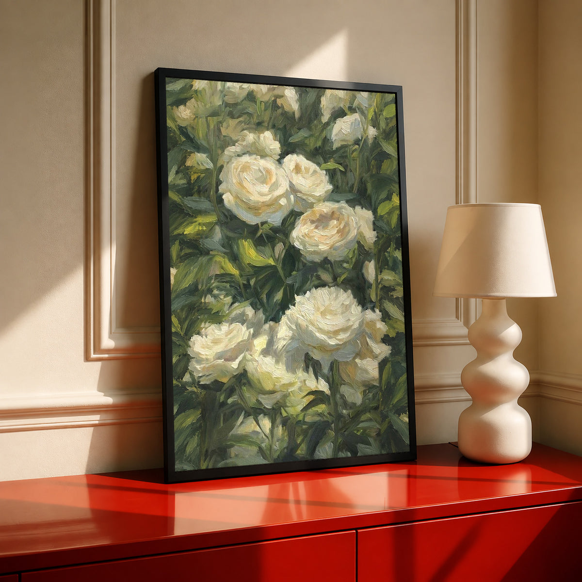 Cream white roses among dense green garden foliage in a painterly composition in a black frame on a red cabinet beside a cream lamp